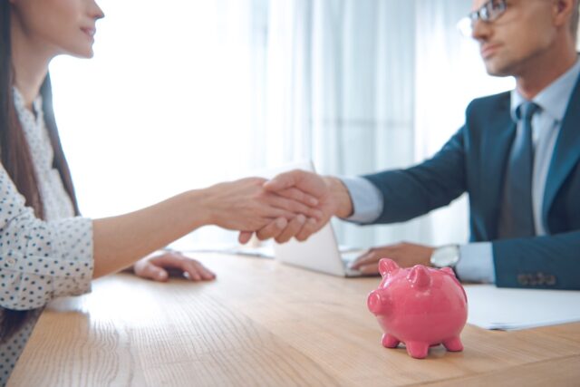 A man and a woman shaking hands across a table with a pink piggy bank and a laptop in the foreground, discussing options for the lowest interest personal loan Singapore has to offer.