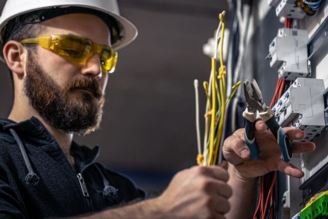 A licensed electrician in Singapore, wearing safety glasses and a hard hat, uses pliers to work on electrical wires inside a control panel.