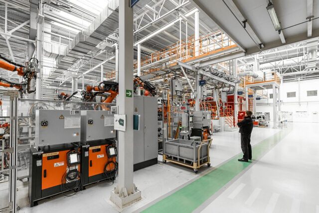 A worker stands in a bright, modern factory with automated machinery, production placement systems, control panels, and robotic arms visible throughout the industrial environment.