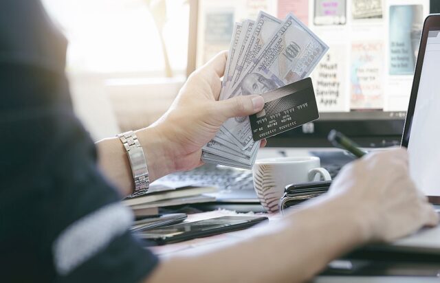 A person holds several US dollar bills and a credit card in one hand while applying for a foreigner loan in Singapore at a desk with a laptop, smartphone, pen, and coffee cup.