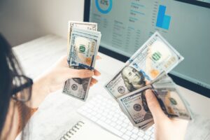 Person holding and counting US $100 bills in front of a computer screen displaying financial graphs and charts, considering options like foreigner loan Singapore to manage international finances.