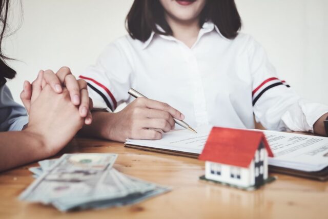 Two women sit at a table with documents, a small house model, and cash, suggesting a financial or real estate transaction, possibly discussing a house reno loan.