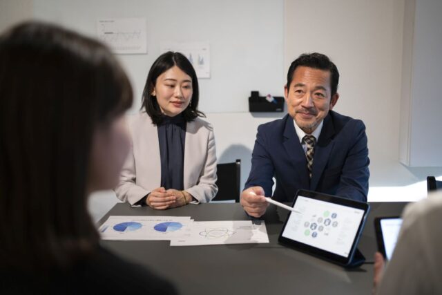 Four people in business attire from a Singapore top audit firm have a meeting around a table with charts and a tablet displaying graphs and data.