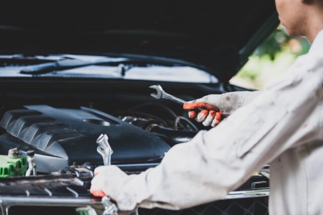 A person wearing gloves holds wrenches while working on a car engine with the hood open, demonstrating effective auto repair tips for better vehicle maintenance.